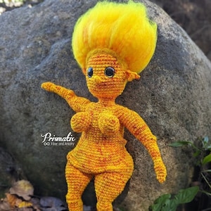May include: A crocheted doll with a bright yellow and orange body and a fluffy yellow hairdo. The doll is standing on a rock with green moss and brown leaves around it.