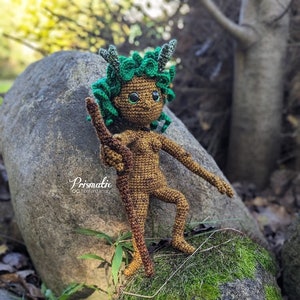 May include: A crocheted woodland creature doll with green hair, brown skin, and a stick in its hand. The doll is standing on a mossy rock.