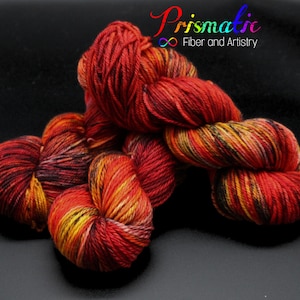 May include: A skein of red, orange, yellow, and black yarn with a speckled pattern. The yarn is arranged in a loose pile on a black surface. The text "Prismatic Fiber and Artistry" is visible in the upper right corner of the image.