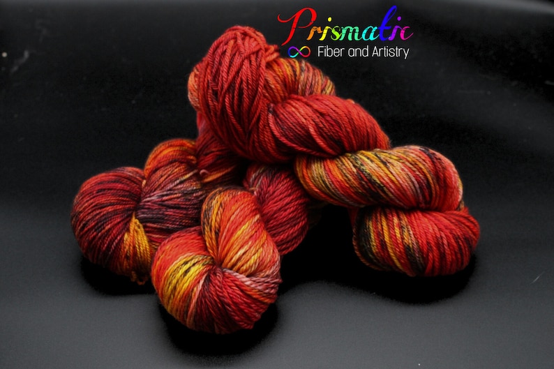 May include: A skein of red, orange, yellow, and black yarn with a speckled pattern. The yarn is arranged in a loose pile on a black surface. The text "Prismatic Fiber and Artistry" is visible in the upper right corner of the image.
