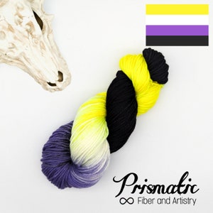 Könnte beinhalten: Eine Garnrolle in den Farben Gelb, Lila und Schwarz. Das Garn ist zu einer lockeren Spirale angeordnet. Das Garn ist mit "Prismatic Fiber and Artistry" beschriftet.