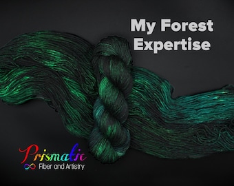 May include: A skein of black and green yarn with a shimmery finish. The yarn is arranged in a loose loop and the text "My Forest Expertise" is visible above the yarn. The text "Prismatic Fiber and Artistry" is visible below the yarn.