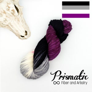 Könnte beinhalten: Eine Garnrolle in den Farben Schwarz, Grau, Weiß und Lila. Das Garn ist zu einer lockeren Spirale angeordnet. Das Garn ist mit "Prismatic Fiber and Artistry" beschriftet.