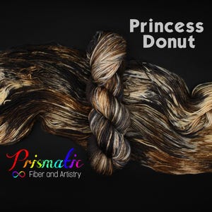 Può includere: Una matassa di filato tinto a mano con una tavolozza di colori variegati in nero, marrone scuro e crema. Il filato è mostrato in una matassa attorcigliata e in una forma sciolta e distesa. Il testo Princess Donut e Prismatic Fiber and Artistry è visibile.