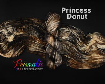 May include: A skein of hand-dyed yarn featuring a variegated color palette of black, dark brown, and cream. The yarn is displayed in a twisted hank and a loose, spread-out form. The text Princess Donut and Prismatic Fiber and Artistry are visible.