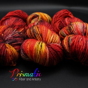 May include: Close-up of six skeins of yarn in a fiery red, orange, and yellow colorway. The yarn is arranged in a pile, with the skeins overlapping each other. The yarn is soft and fluffy, and the colors are vibrant and rich.