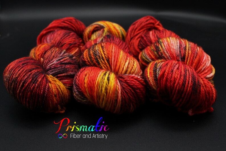 May include: Close-up of six skeins of yarn in a fiery red, orange, and yellow colorway. The yarn is arranged in a pile, with the skeins overlapping each other. The yarn is soft and fluffy, and the colors are vibrant and rich.