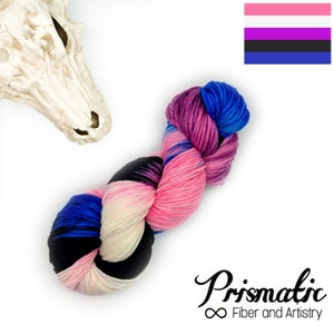 Könnte beinhalten: Eine Garnrolle in den Farben Rosa, Lila, Blau, Schwarz und Weiß. Das Garn ist in einer lockeren Schlaufe angeordnet und trägt die Aufschrift "Prismatic Fiber and Artistry".