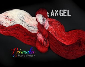 May include: A skein of yarn in a red, white, and black colorway. The yarn is arranged in a loose loop and the text "ANGEL" is visible in the background. The text "Prismatic Fiber and Artistry" is visible in the lower left corner.