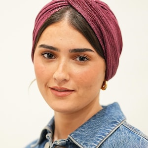 Peut inclure: Une femme porte un turban bordeaux avec une finition texturée et chatoyante. Le turban est noué sur le dessus et est associé à une veste en jean et des créoles dorées. Le fond est blanc uni.