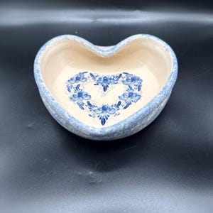 May include: A heart-shaped ceramic bowl with a cream-colored interior featuring a blue floral pattern arranged in a heart shape. The exterior rim and sides have a mottled blue and white sponge-painted finish, creating a textured appearance.