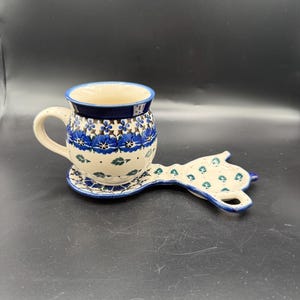May include: A ceramic mug and spoon rest set. The mug has a cream-colored interior and is decorated with blue floral patterns. The spoon rest, shaped like a cat, mirrors the mug's design. The set is on a dark surface.