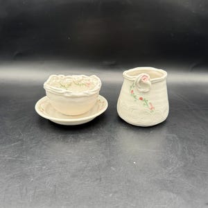 May include: A set of two handmade ceramic pieces with a delicate floral design. The larger piece is a vase with a rounded shape and a small handle. The smaller piece is a bowl with a scalloped edge and a matching saucer.