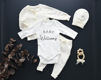 May include: A white baby outfit set with a long-sleeved onesie, pants, cardigan, and hat. The onesie has the text "BABY Williams" printed on the front. The set is shown on a dark blue background. Color shown: Ivory.