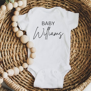 May include: White baby onesie with black text that reads "BABY Williams" in a cursive font. The onesie is lying in a woven basket with wooden beads.