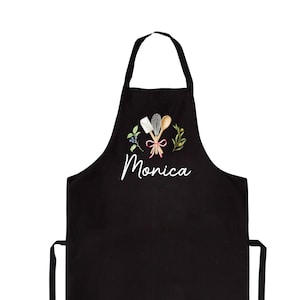 Pu&ograve; includere: Grembiule da cucina nero con un design ricamato bianco di utensili da cucina, un nastro rosso e foglie verdi. Il nome "Monica" &egrave; ricamato sotto il design.