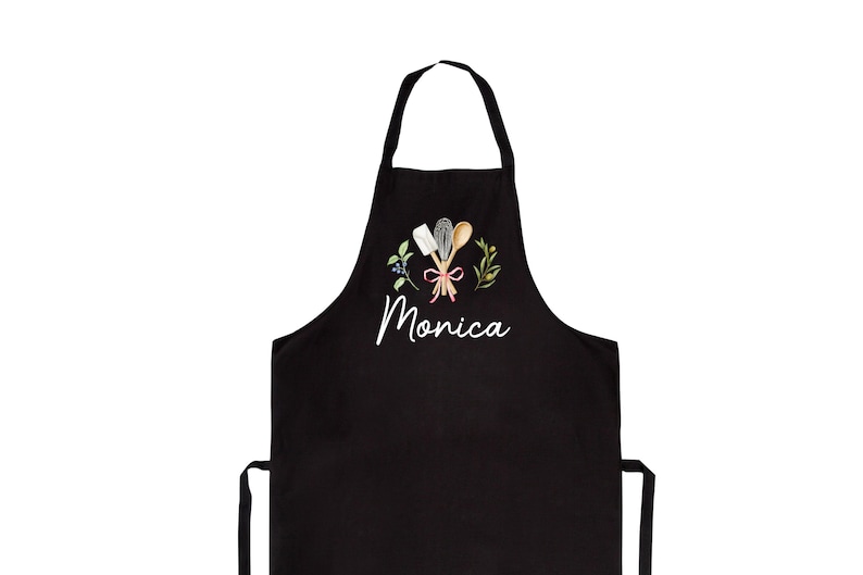 Pu&ograve; includere: Grembiule da cucina nero con un design ricamato bianco di utensili da cucina, un nastro rosso e foglie verdi. Il nome "Monica" &egrave; ricamato sotto il design.