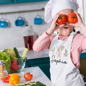 Pu&ograve; includere: Una bambina con un cappello da cuoco bianco e un grembiule bianco con il nome "Sophia" ricamato. Sta tenendo due pomodori rossi davanti ai suoi occhi, sorridendo.