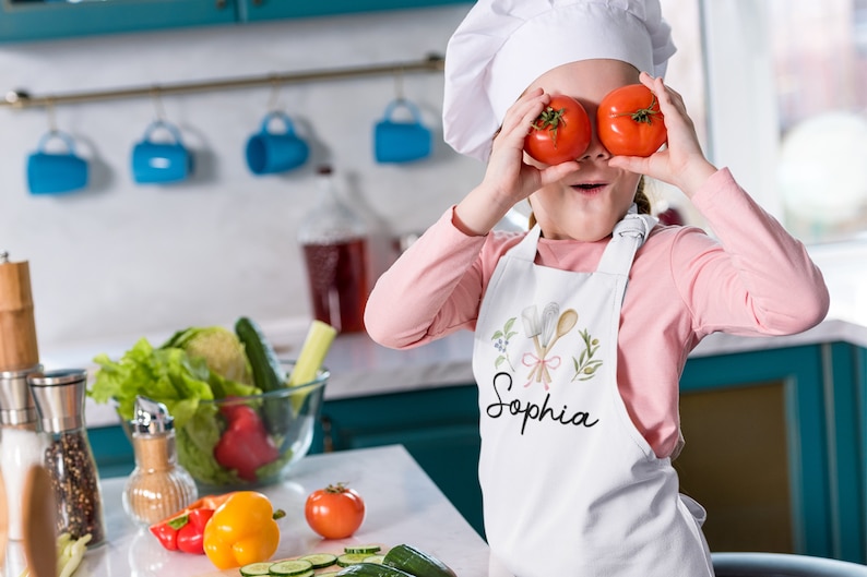 Pu&ograve; includere: Una bambina con un cappello da cuoco bianco e un grembiule bianco con il nome "Sophia" ricamato. Sta tenendo due pomodori rossi davanti ai suoi occhi, sorridendo.