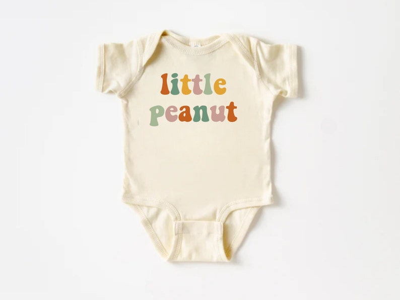 May include: A cream-colored baby onesie with the text "little peanut" in colorful retro-style lettering.