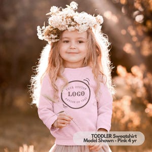 May include: Pink toddler sweatshirt with a circle design that says "Your Custom Logo Design or Text". The model is wearing a flower crown and a pink skirt.