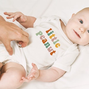 May include: A white baby onesie with the text "little peanut" in rainbow colors. The baby is wearing the onesie and is lying on a white surface.