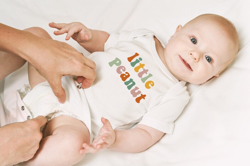 May include: A white baby onesie with the text "little peanut" in rainbow colors. The baby is wearing the onesie and is lying on a white surface.