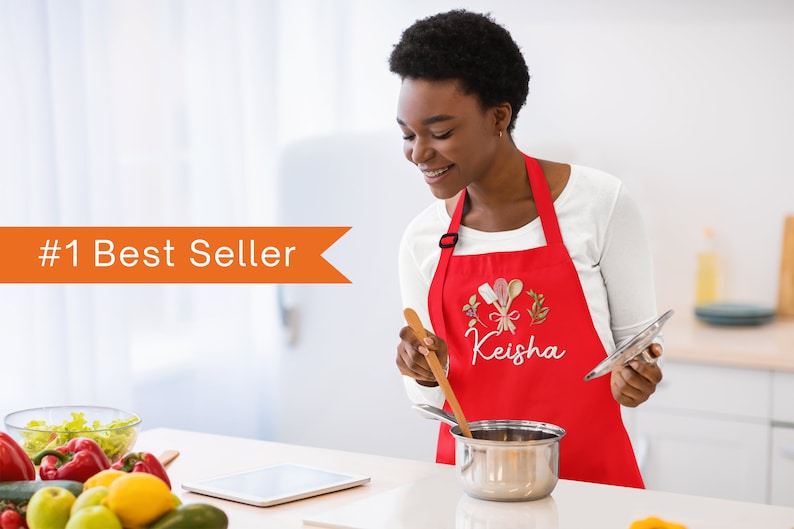 Pu&ograve; includere: Un grembiule rosso personalizzato con un design ricamato bianco di utensili da cucina e il nome "Keisha".