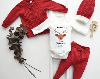 Può includere: Un cardigan di lana rosso, un body bianco a maniche lunghe con un'immagine di renna e il testo "Emma's First Christmas 2024", pantaloni di lana rossi e un cappello di lana rosso con un pon pon bianco.