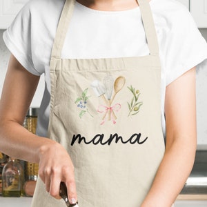 Pu&ograve; includere: Un grembiule da cucina beige con un design ad acquerello di utensili da cucina, vegetazione e bacche. Il grembiule ha la parola "mama" stampata in nero sotto il design.