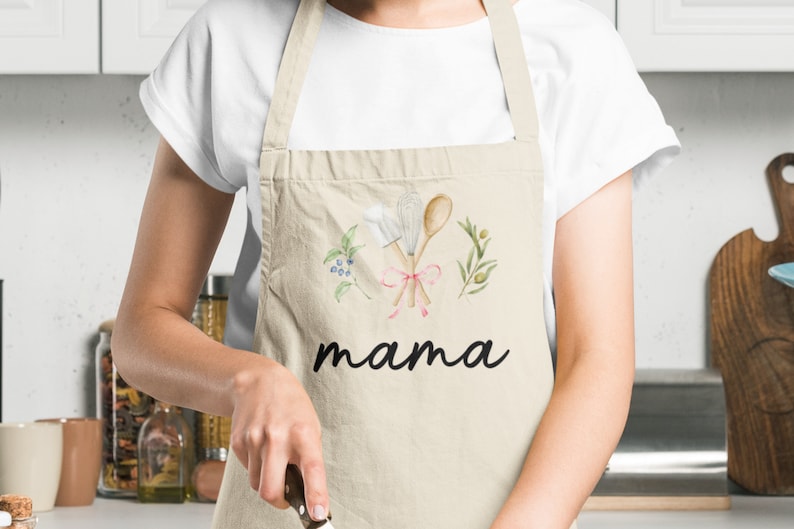 Pu&ograve; includere: Un grembiule da cucina beige con un design ad acquerello di utensili da cucina, vegetazione e bacche. Il grembiule ha la parola "mama" stampata in nero sotto il design.