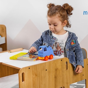 May include: A young child sits at a wooden table with a white surface. The table has a built-in chair and a shelf for books. The child is playing with a blue toy truck and a book. The table is made by the company "minera".