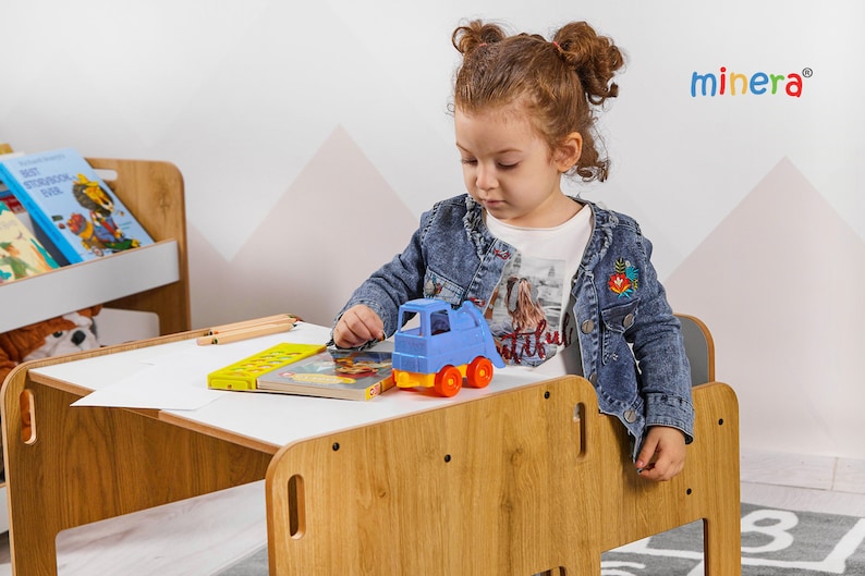 May include: A young child sits at a wooden table with a white surface. The table has a built-in chair and a shelf for books. The child is playing with a blue toy truck and a book. The table is made by the company "minera".