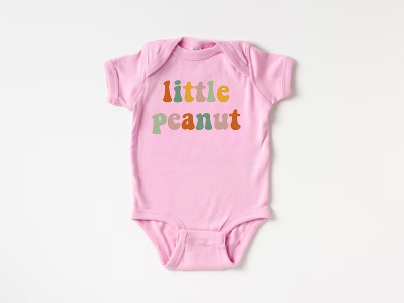 May include: Pink baby onesie with the text "little peanut" in colorful, retro-style letters.