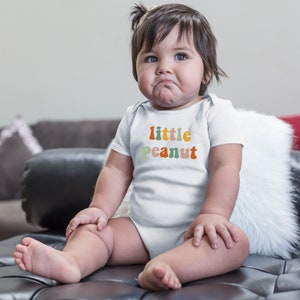 May include: A white baby onesie with the text "little peanut" in colorful, retro-style letters.