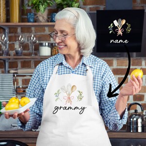 Pu&ograve; includere: Un grembiule bianco con un disegno floreale e la parola "grammy" stampata su di esso. Il grembiule presenta un disegno di cucchiai di legno incrociati con un nastro rosa e foglie verdi. Il grembiule &egrave; indossato da una persona in un ambiente da cucina.