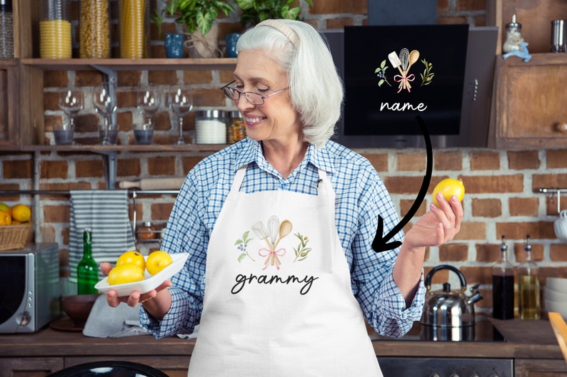 Pu&ograve; includere: Un grembiule bianco con un disegno floreale e la parola "grammy" stampata su di esso. Il grembiule presenta un disegno di cucchiai di legno incrociati con un nastro rosa e foglie verdi. Il grembiule &egrave; indossato da una persona in un ambiente da cucina.