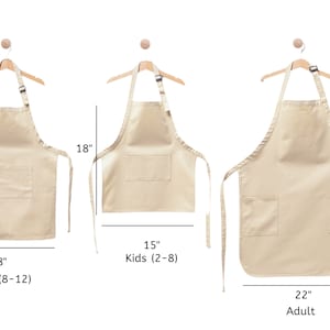 Pu&ograve; includere: Tre grembiuli in cotone beige di diverse dimensioni, appesi a grucce di legno. I grembiuli hanno una sola tasca e lacci regolabili. Le taglie sono indicate come Giovani (8-12), Bambini (2-8) e Adulti, con le misure corrispondenti in pollici.