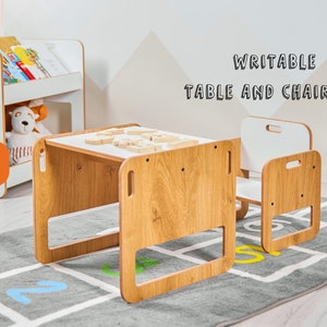 May include: A white and wood children's table and chair set with a white top that can be written on. The set is on a gray rug with numbers printed on it. The text "WRITEABLE TABLE AND CHAIR SET" is printed on the wall behind the set.