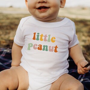 May include: A white baby onesie with the text "little peanut" in colorful, retro-style lettering. The onesie is worn by a baby sitting on a plaid blanket.