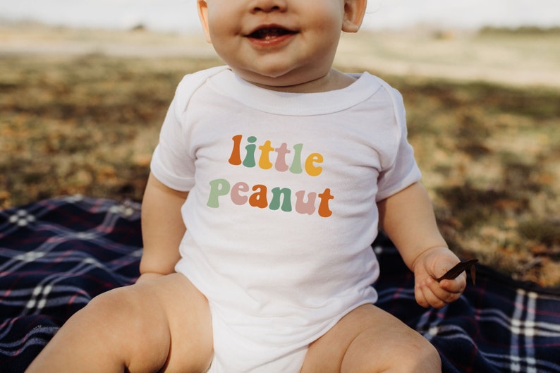 May include: A white baby onesie with the text "little peanut" in colorful, retro-style lettering. The onesie is worn by a baby sitting on a plaid blanket.