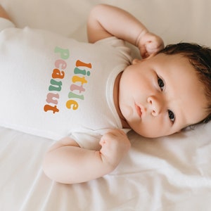 May include: A baby wearing a white onesie with the text "little peanut" in colorful letters. The baby is lying on a white sheet and has their arm raised above their head.
