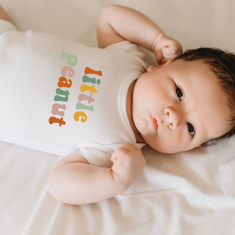 May include: A baby wearing a white onesie with the text "little peanut" in colorful letters. The baby is lying on a white sheet and has their arm raised above their head.