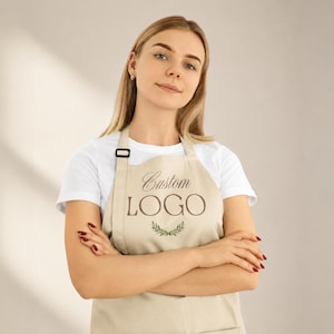Pode incluir: Um avental bege com o texto Custom LOGO em letras castanhas, acompanhado por um pequeno gráfico de folhas verdes centrado abaixo do texto. O avental tem uma fivela preta ajustável na alça do pescoço e é usado sobre uma t-shirt branca.
