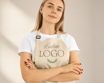May include: A beige apron featuring the text Custom LOGO in brown lettering, accompanied by a small green leaf graphic centred below the text. The apron has a black adjustable neck strap buckle and is worn over a plain white short-sleeved t-shirt.