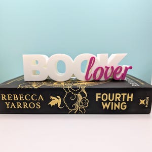 Peut inclure: Un livre noir avec des lettres dorées et un signe "Book Lover" blanc et magenta sur le dessus. Le titre du livre est "Fourth Wing" de Rebecca Yarros.