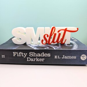 May include: A white 3D-printed word art piece that reads "SM slut" in a bold font, with the word "slut" in red. The piece is placed on top of a book titled "Fifty Shades Darker" by EL James.