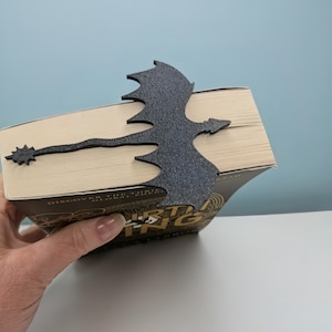 May include: A black dragon-shaped bookmark made of textured material. The dragon is positioned over the pages of a book, with its tail extending to the left. The book has a gold and black cover.