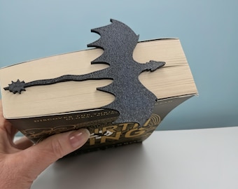 Puede incluir: Un marcapáginas negro con forma de dragón hecho de material texturizado. El dragón está colocado sobre las páginas de un libro, con su cola extendiéndose hacia la izquierda. El libro tiene una cubierta dorada y negra.