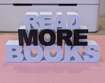 Può includere: Un'insegna decorativa con la scritta "READ MORE BOOKS" in lettere a blocchi 3D. Le parole "READ" e "BOOKS" sono bianche, mentre "MORE" è nera. L'insegna è su una superficie rosa, con una stampante bianca sullo sfondo.